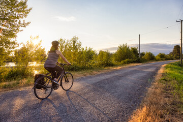 Adventurous White Cacasusian Woman riding a bicycle on a road. Sunny Summer Sunset. Barnston Island, Vancouver, British Columbia, Canada. Adventure Journey Concept