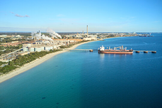 horizontal shot of Kwinana alumina refinery on a sunny day with white smoke, bushes and docked ship