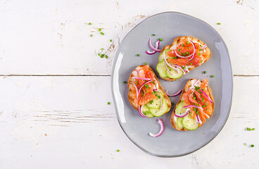 Fototapeta na wymiar Toasts with cream cheese, smoked salmon, cucumber and red onion on rustic wooden table. Open sandwiches. Healthy care, super food concept. Top view, overhead