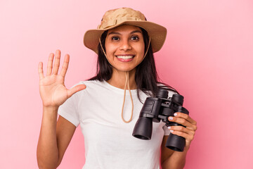 Young latin woman holding binoculars isolated on pink background smiling cheerful showing number five with fingers.