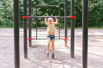 little girl pulls herself up on a horizontal bar in a city park on a summer day