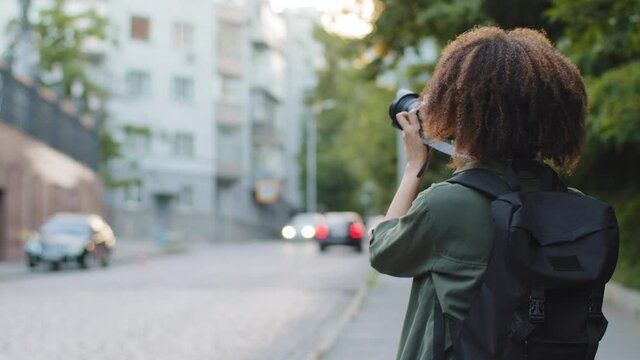 Curly-haired girl tourist with backpack walking along road photographing attractions in big city. Young woman photographer blogger sightseeing takes pictures using professional camera during travel