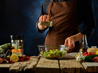 The chef spices the ingredients of Greek salad in a large glass salad bowl that stands on a plain wooden table. All the ingredients for the salad are next to it. Dark blue background. Close-up.