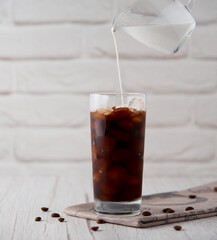 Iced coffee in a tall glass with cream on a white wood background. Cold tasty summer refreshment beverage concept. Selective focus, copyspace.