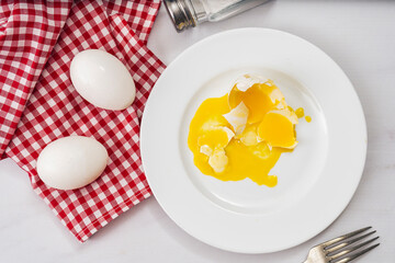 Aerial view of a broken chicken egg on a white plate next to two healthy eggs on a red and white country napkin.