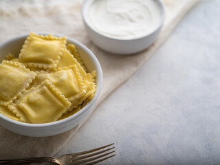 Delicious and healthy ravioli dish. On a white plate, a bowl with delicious ravioli drizzled with butter. There is a bowl of sour cream next to it. There is a fork near the bowl. High angle view.