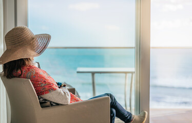 Close-up of a woman seated, gazing out of a hotel room window at the sea.