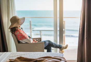 a woman sitting, gazing at the sea through a window from a hotel room.