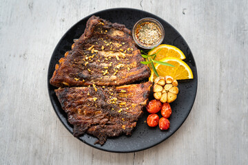 A smoked pork spare ribs BBQ steak with organic ingredient such as garlic, tomato and lemon is served in black plate, placed on the white wooden table. Food photo, close-up and selective focus.