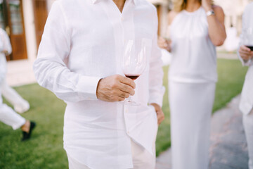Man in a white shirt and trousers stands with a glass of red wine in his hand