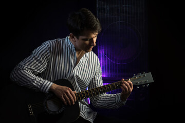 Obraz premium Photo of man sitting and playing acoustic guitar in recording studio on dark background with light effect. Repetition of the rock music of the guitarist. Rehearsal base