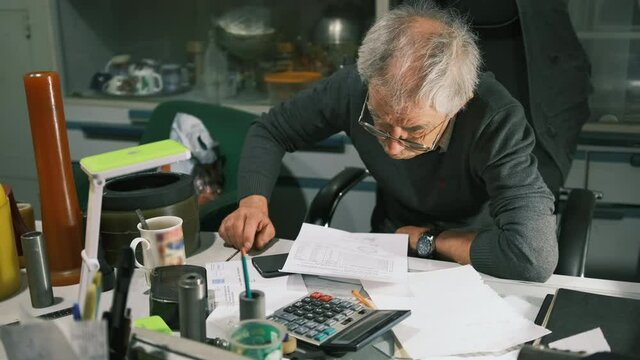 An elderly man sits at his desk and examines a document and is very surprised. Elderly man looks at the bill is stunned and shocked