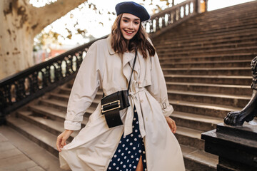 Trendy young lady with brunette hair in beret, beige trench coat and crossbody bag, smiling and...