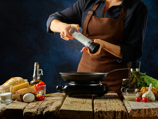 The chef prepares a classic Caesar salad. There is a frying pan on a wooden table. The cook will salt the ingredients that are fried in the pan. A lot of salad products are nearby.