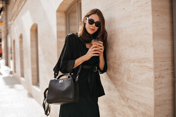 Gorgeous young woman with brunette wavy hairstyle, sunglasses, black coat and bag walking in daylight city and posing against beige wall background