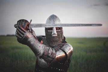 Antique knight in the plate armor and helmet holds a sword in hands close up. - obrazy, fototapety, plakaty
