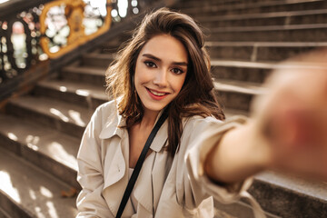 Beautiful young brunette in trench beige trench coat sitting on stairs outdoors, smiling, making selfie and looking into camera. Warm sunny autumn