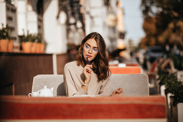 Charming young lady with brunette wavy hairstyle, red lips and stylish glasses, beige pullover,...