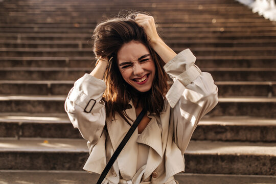 Joyful young brunette with beige trench coat posing, closing eyes, shaking hair by hands and having fun against old city stairs background outdoors