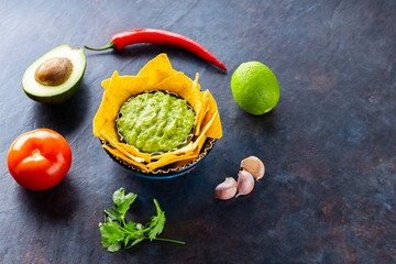 Guacamole sauce with ingredients avocado, pepper, lime and corn nachos on a dark background. Guacamole with ingredients and nachos tortilla chips. Copy space