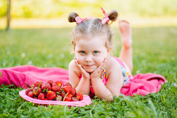 Adorable little girl lie on the grass with plate of fresh ripe strawberries and smile. Childhood, immunity, happiness.