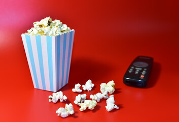 Popcorn in colored cardboard packaging with TV remote for listening to music and watching a movie. Heap of salted popcorn in paper striped bucketon a red background.