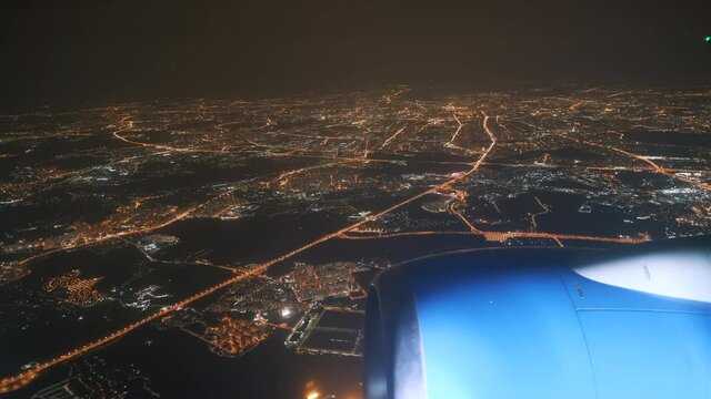 View from the plane window of the lights of the glowing city at night and the plane's engine on the wing. Flight over the metropolis at night on an airplane view from the wing window.