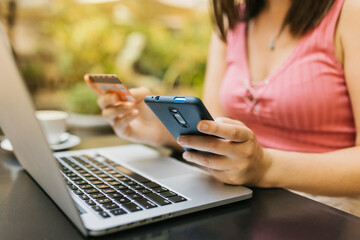 Close up woman hands holding credit card while using laptop and mobile phone for online shopping at restaurant cafe. Female buyer texting her secret number on smartphone bank app while buying and