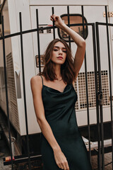 Fototapeta na wymiar Gorgeous young brunette with bright makeup, well-built body, silk long slip dress, posing on street, raising one hand and looking into camera