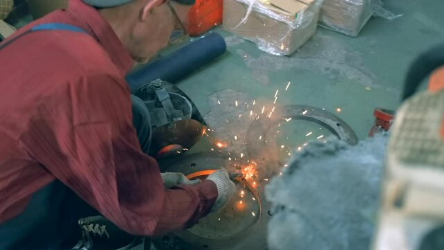 Side view An elderly male blacksmith welder works without a protective mask in glasses, welds metal parts with a welding machine. Sparks and flashes from the welding machine.