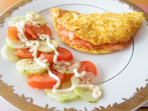 An omelet with a smaked salmon filling on a white plate with a gold trim. There is also a tomato and cumcumber salad on the plate.