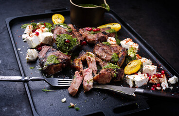 Traditional barbecue T-Bone lamb steaks with Greek feta cheese, chimichurri sauce and pomegranate fruit served as close-up on a black design tray