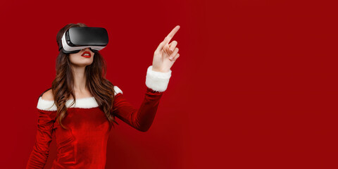 Amazed young Santa girl in Christmas dress looking in headset pointing hands aside isolated on red wall background. 