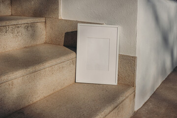 Blank white picture frame leaning against white wall. Outdoor sandstone stairs in sunlight, shadows...