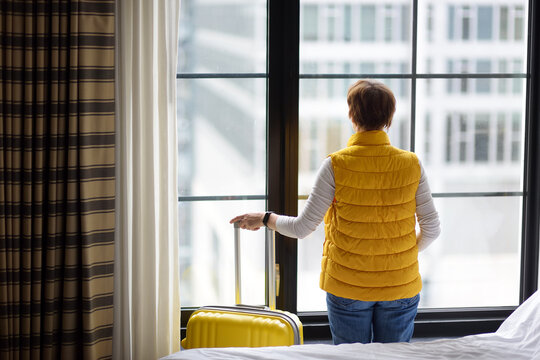 Woman tourist stays in hotel room in New York. Traveler with suitcase looks and admires of view the skyscrapers of Manhattan outside the window. Tourism and travel in USA. Booking apartment
