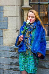 Dressing in a blue faux fur plus size jacket, patterned dress and scarf, holding a white flower, a pretty girl is standing by a window in winter, smelling, looking away and thinking..
