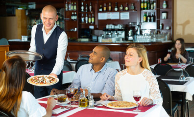 Fototapeta na wymiar Man waiter is brings pizza to friends who have dinner and drink wine in restaurant