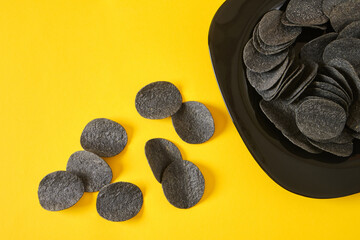 black potato chips on a black plate, yellow background, copy space, top view