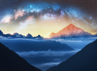 Milky Way arch and snowy mountains in low clouds at starry night. Landscape. Sky with stars, arched...