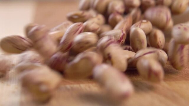 Dry uncooked cranberry beans falling from a white bowl onto a wooden cutting board in slow motion. Macro shot