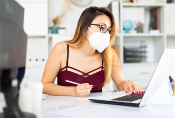 Positive latina woman in protective mask and casual wear working alone with laptop and papers in office