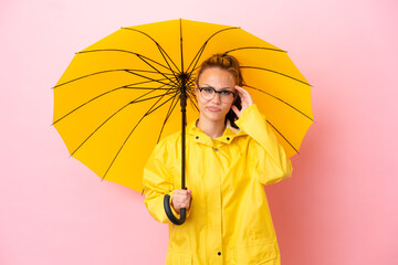 Teenager Russian girl with rainproof coat and umbrella isolated on pink background having doubts