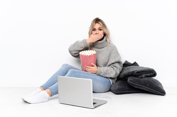 Teenager blonde girl eating popcorn while watching a movie on the laptop covering mouth with hands