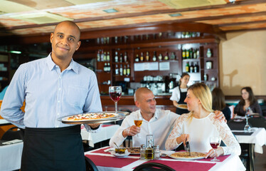 Fototapeta na wymiar Portrait of friendly Hispanic waiter holding plate with appetizing pizza, warmly welcoming in cozy pizzeria