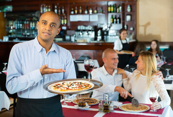 Hospitable latin american waiter standing with serving tray in pizza restaurant hall, welcoming guests
