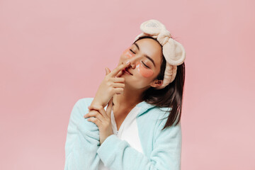 Brunette pretty woman in blue pajamas and soft beige headband touches nose. Cute Asian girl in sleep wear poses with eyepatches on pink background.