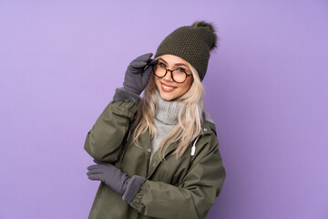 Teenager blonde girl with winter hat over isolated purple background with glasses and happy