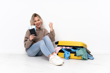 Teenager blonde girl with a suitcase full of clothes sitting on the floor celebrating a victory