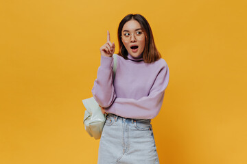 Teen cool brunette Asian woman in eyeglasses, purple sweater and denim skirt points up, has great new idea and poses with backpack on orange background.