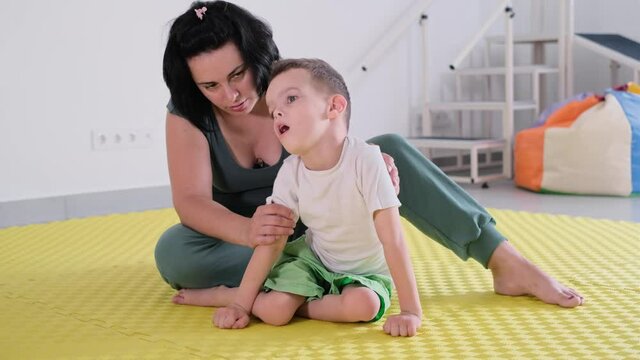 Therapist doing exercises on the mat with disabled child. Boy with cerebral palsy having rehabilitation. Spine and neck, foots training on the floor of center. Learning to sit while playing